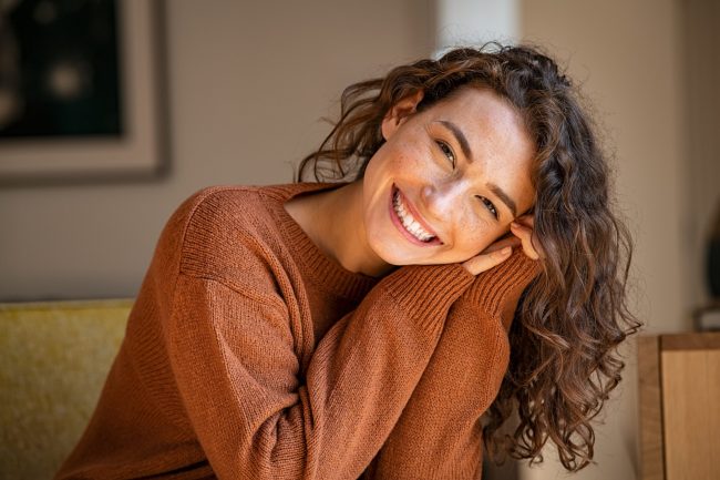 Woman leaning on her arms on table in brown sweater looking happy with glowing skin after utilizing holistic skincare practices